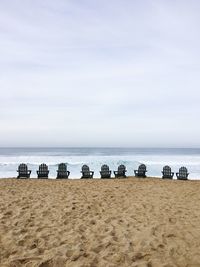 Empty adirondack chairs at beach against sky