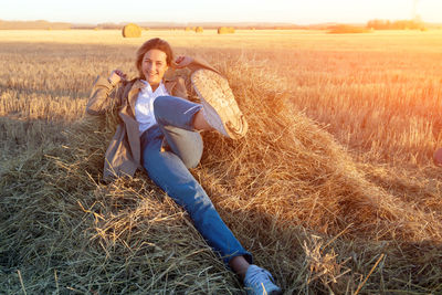 A young woman in a white shirt, beige coat and jeans enjoys nature, sitting on a haystack 