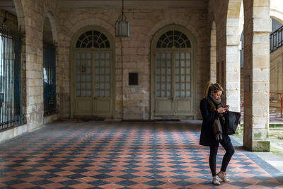 Full length of young woman using mobile phone while standing in corridor