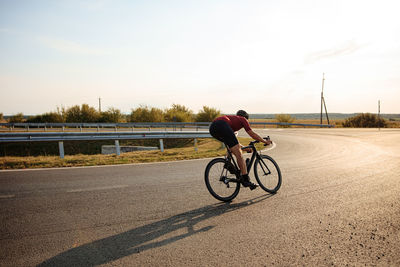 Muscular athlete dressed in sport outfit cycling on paved road during morning sun. 