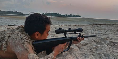 Side view of shirtless young man aiming rifle while lying on sand against sky during sunset