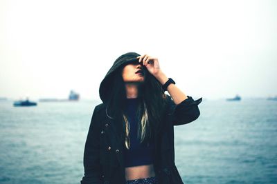 Beautiful young woman in hooded jacket standing against sea