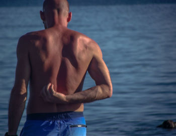 Rear view of shirtless man scratching while standing against sea