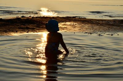 Rear view of silhouette girl sitting in water at beach during sunset