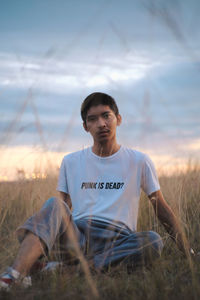 Portrait of young man sitting on grassy land against sky at sunset