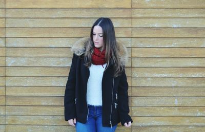 Thoughtful woman wearing fur coat while standing against wooden wall