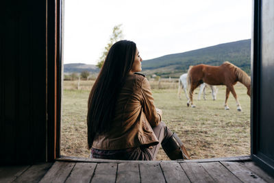 Back view of stylish woman in boots and scarf sitting on porch of old wooden house in countryside looking away