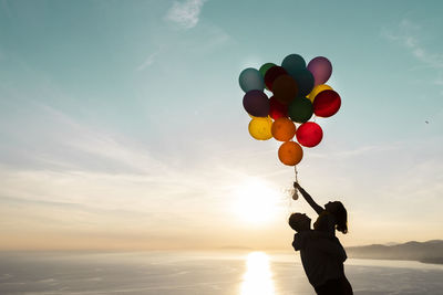 Low angle view of woman holding colorful balloons against sky during sunset