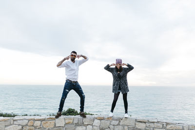 Full length of couple gesturing while standing on retaining wall against sea