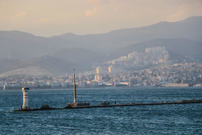 Lighthouse in sea by city and mountains against sky