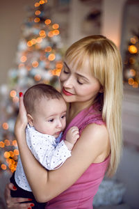 Woman in a pink dress holds her son's child in her arms standing at the christmas tree in the studio