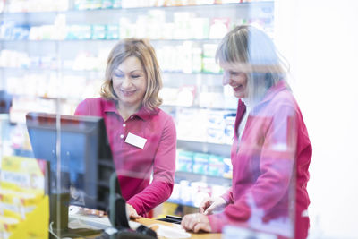 Happy pharmacist using computer and talking with colleague seen through glass