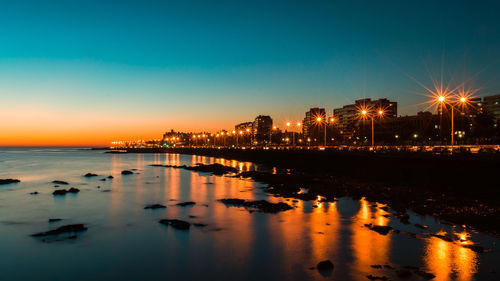 Illuminated city by sea against sky at sunset