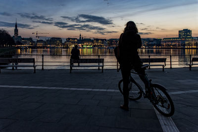 Man with bicycle on river in city at sunset
