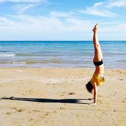 Side view of woman doing handstand at beach against sky