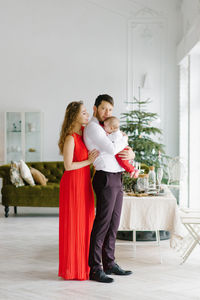 A beautiful family with a baby in their arms stand near the window in the living room, decorated 