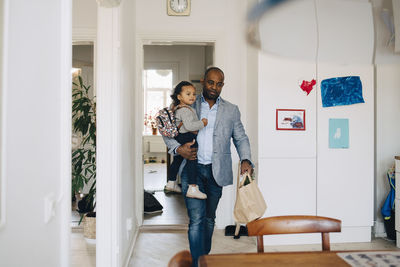 Father carrying daughter with backpack while walking in living room at home