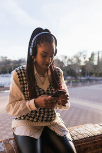 Charming african american female with braids sitting on stone fence on plaza de espana and enjoying music in headphones while browsing smartphone