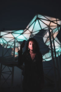 Portrait of woman holding mobile phone while standing against illuminated lights at night