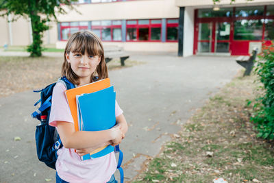 Portrait of a schoolgirl with a backpack on her back and textbooks in her hands against the school.