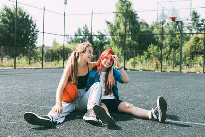 Two girls in sports clothes and with a basketball are chatting, sitting on the playground. 