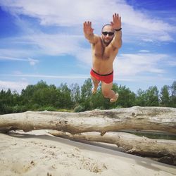 Shirtless man jumping over logs at beach against sky