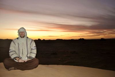 Portrait of man sitting on beach against sky during sunset