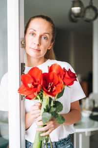 Portrait of young woman in white t-shirt with bouquet of red flowers in hands near window in kitchen 
