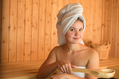 Beautiful young woman holding wooden spoon while sitting in sauna