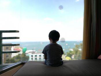 Rear view of boy sitting on bed against glass window