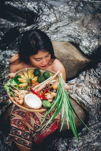 High angle view of woman holding fresh food in bucket while sitting on rock amidst river