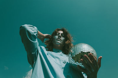 Low angle portrait of woman with glitter on face holding disco ball against clear sky