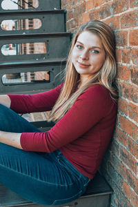 Portrait of beautiful young woman sitting against brick wall