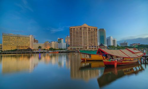 Boats moored in river by buildings against blue sky