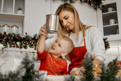 Mother helps her little son in a red sweater sift flour through a sieve in christmas kitchen.