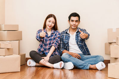 Portrait of couple showing thumbs up while sitting on floor at new home