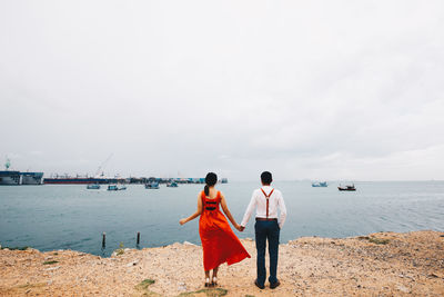 Rear view of couple holding hands while standing at beach against cloudy sky