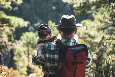  bearded man dressed casual walking in nature and carrying his french bulldog dog on shoulders.