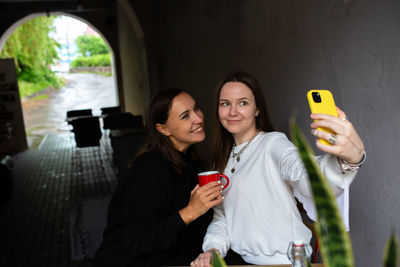 Two women friends have fun outdoors in cafe and take a selfie on a smartphone