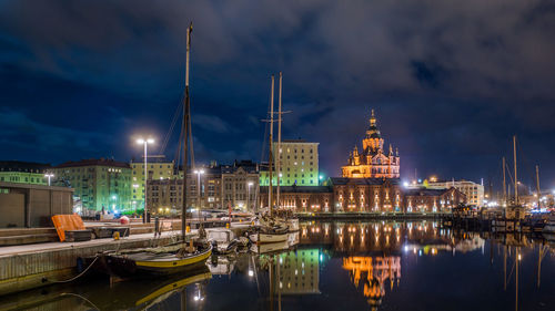 Boats moored at harbor by illuminated buildings against cloudy sky at night