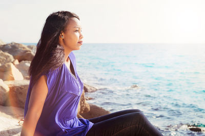 Young woman looking away while sitting on sea shore against sky