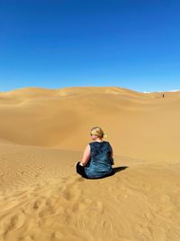 Woman sitting on sand dune in desert against clear sky