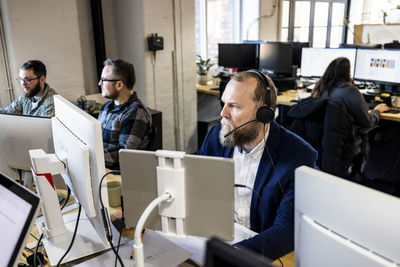 Mature businessman wearing headset working on computer with colleagues at desk