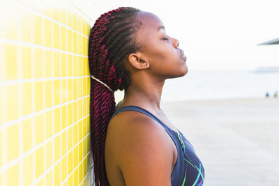 Fit black female athlete in sportswear resting leaning against yellow tiled wall after working out