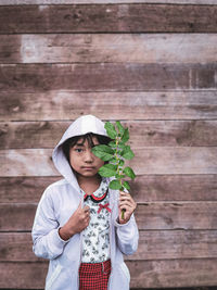 Portrait of girl holding plant standing against wooden wall outdoors