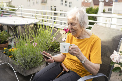 Smiling senior woman holding coffee cup looking at mobile phone on balcony