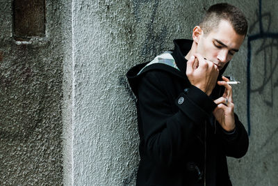 Young man smoking while standing against wall