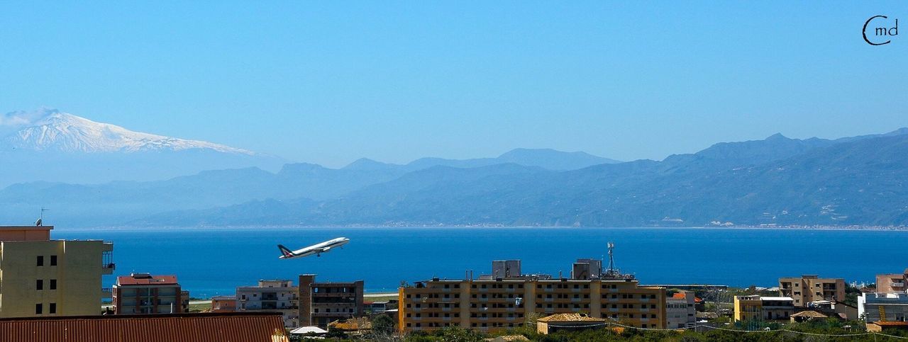 mountain, clear sky, building exterior, blue, mountain range, architecture, built structure, water, copy space, sea, scenics, city, cityscape, nature, transportation, waterfront, beauty in nature, day, outdoors, tranquil scene