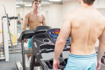 Shirtless young man on treadmill reflecting on mirror in gym