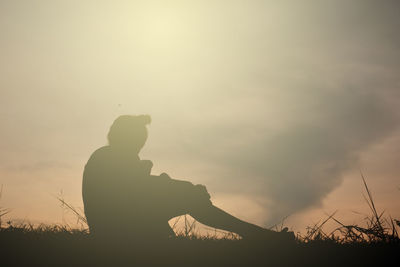 Silhouette girl with teddy bear sitting on field against sky during sunset